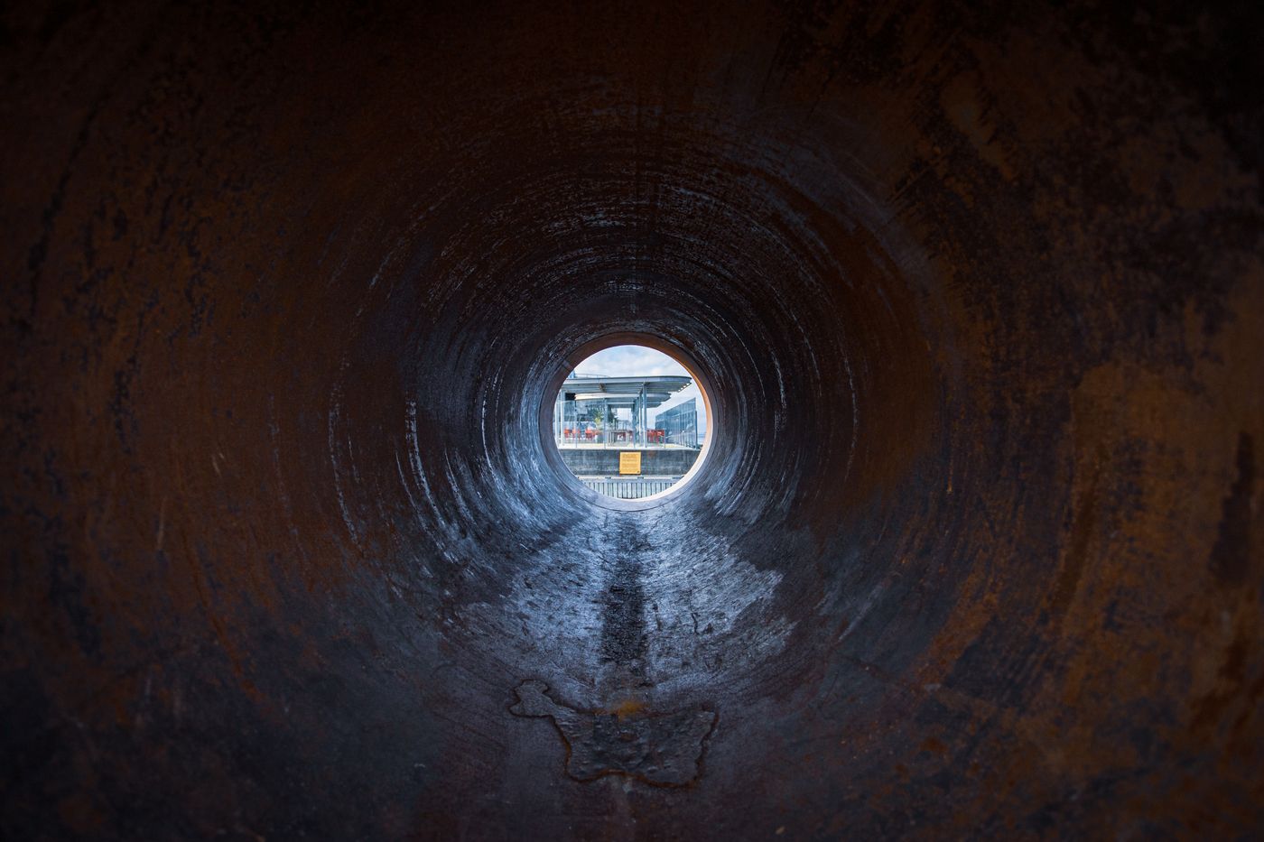 View down the rusty interior of a large pipe toward a bright opening, similar to what a drain inspection camera sees