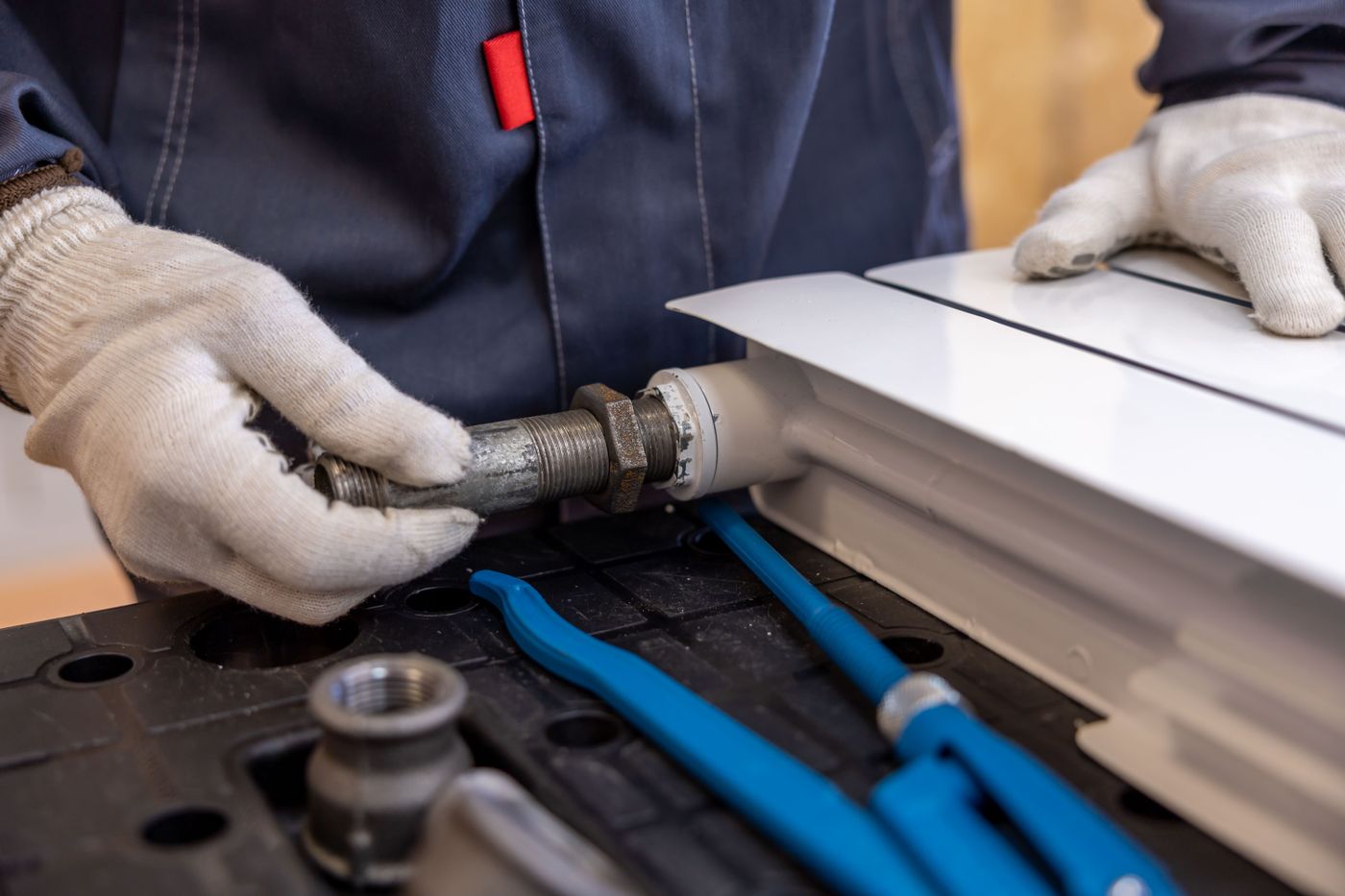 Gloved plumber tightening a threaded pipe fitting with pipe wrenches and fittings laid out nearby