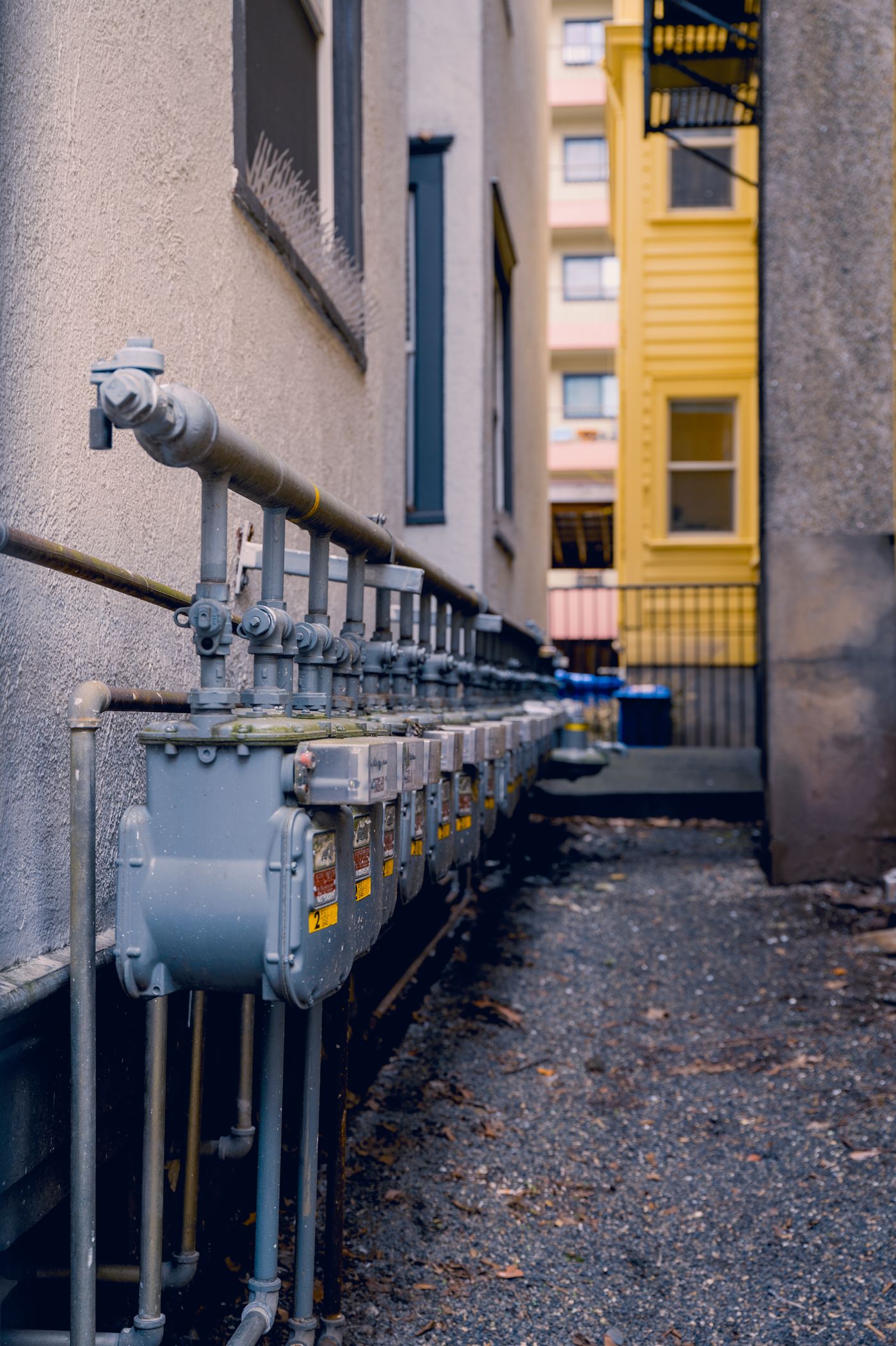 A row of residential gas meters and supply piping mounted along the exterior wall of a building
