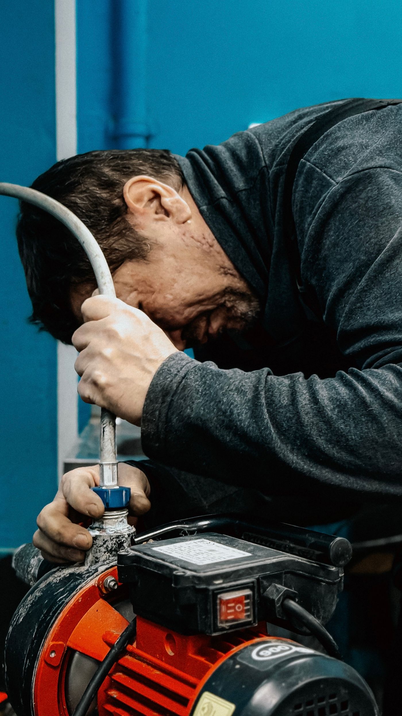 A worker leaning in close to inspect and work on a water pump and connected hose in a utility space