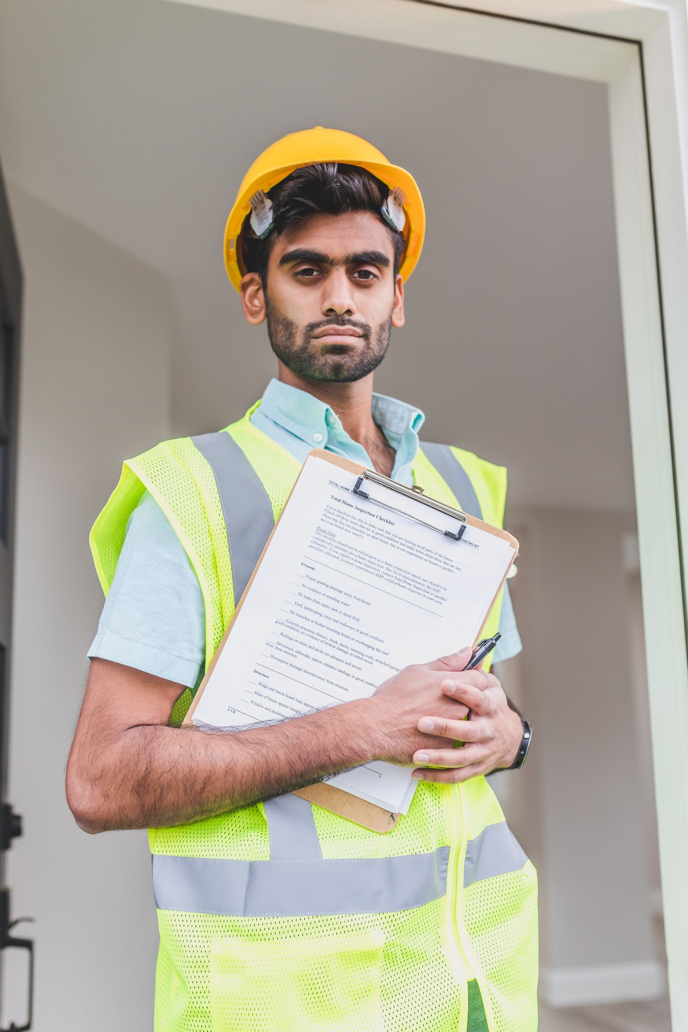 A worker in a hard hat and hi-vis vest holding a clipboard with a home inspection checklist in a doorway