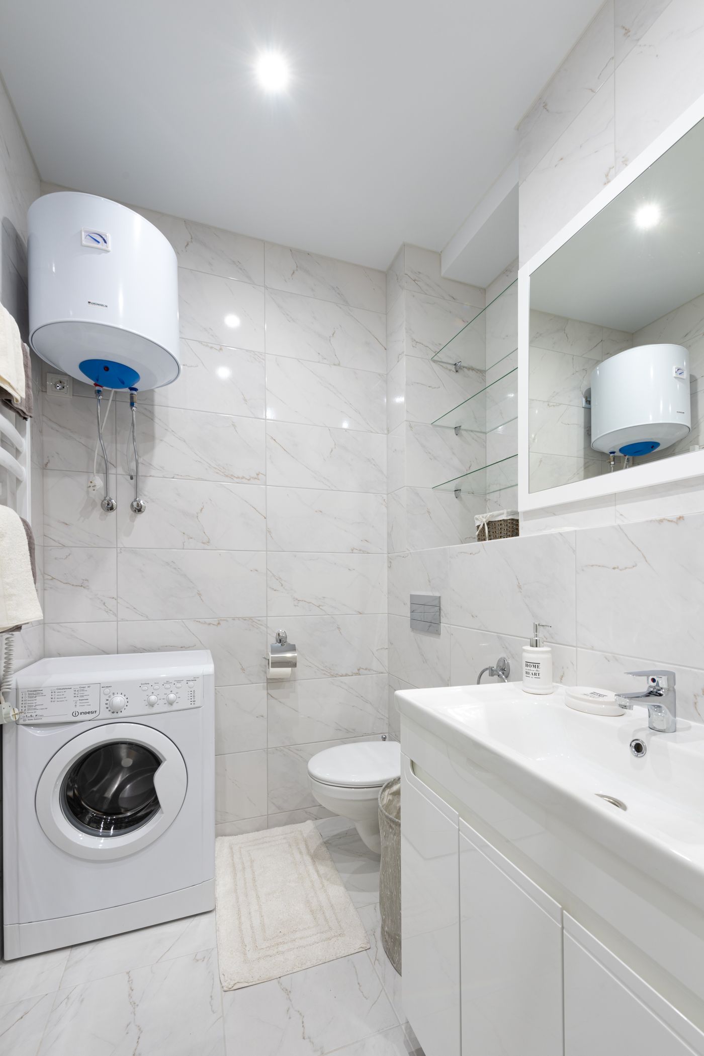 A compact wall-mounted water heater in a small tiled bathroom that also holds a washing machine and sink