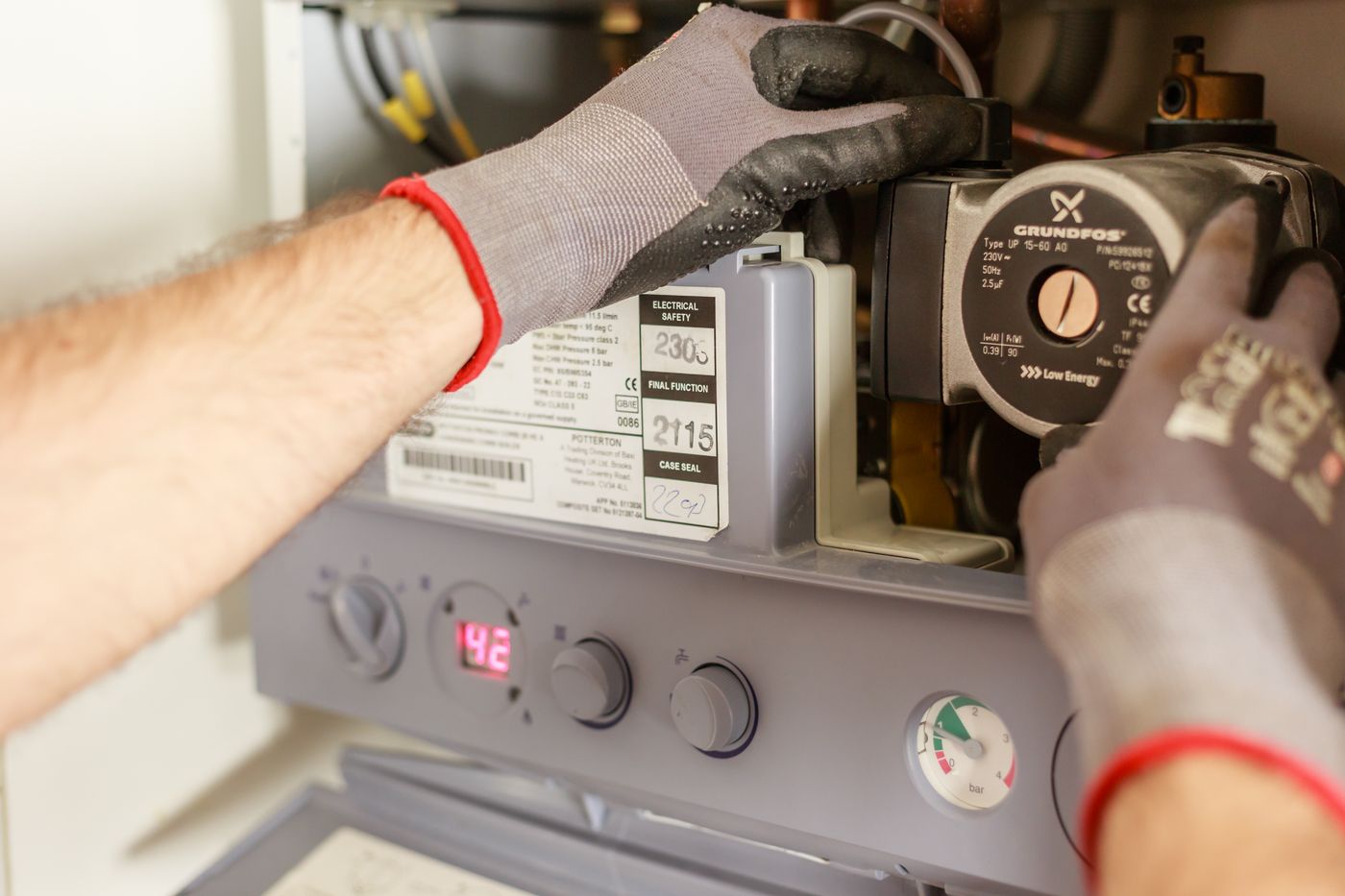 Gloved hands of a technician servicing the internal pump and pressure gauge of a wall-mounted water heater, panel open