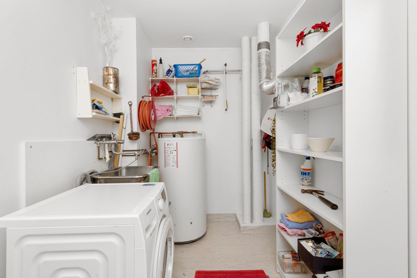 A traditional white cylindrical storage water heater tank standing in a bright utility and laundry room