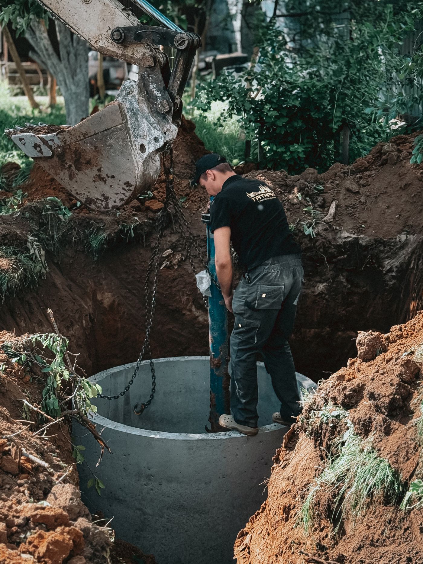 Workers installing a large concrete pipe section into an excavated hole while an excavator lowers it on a chain