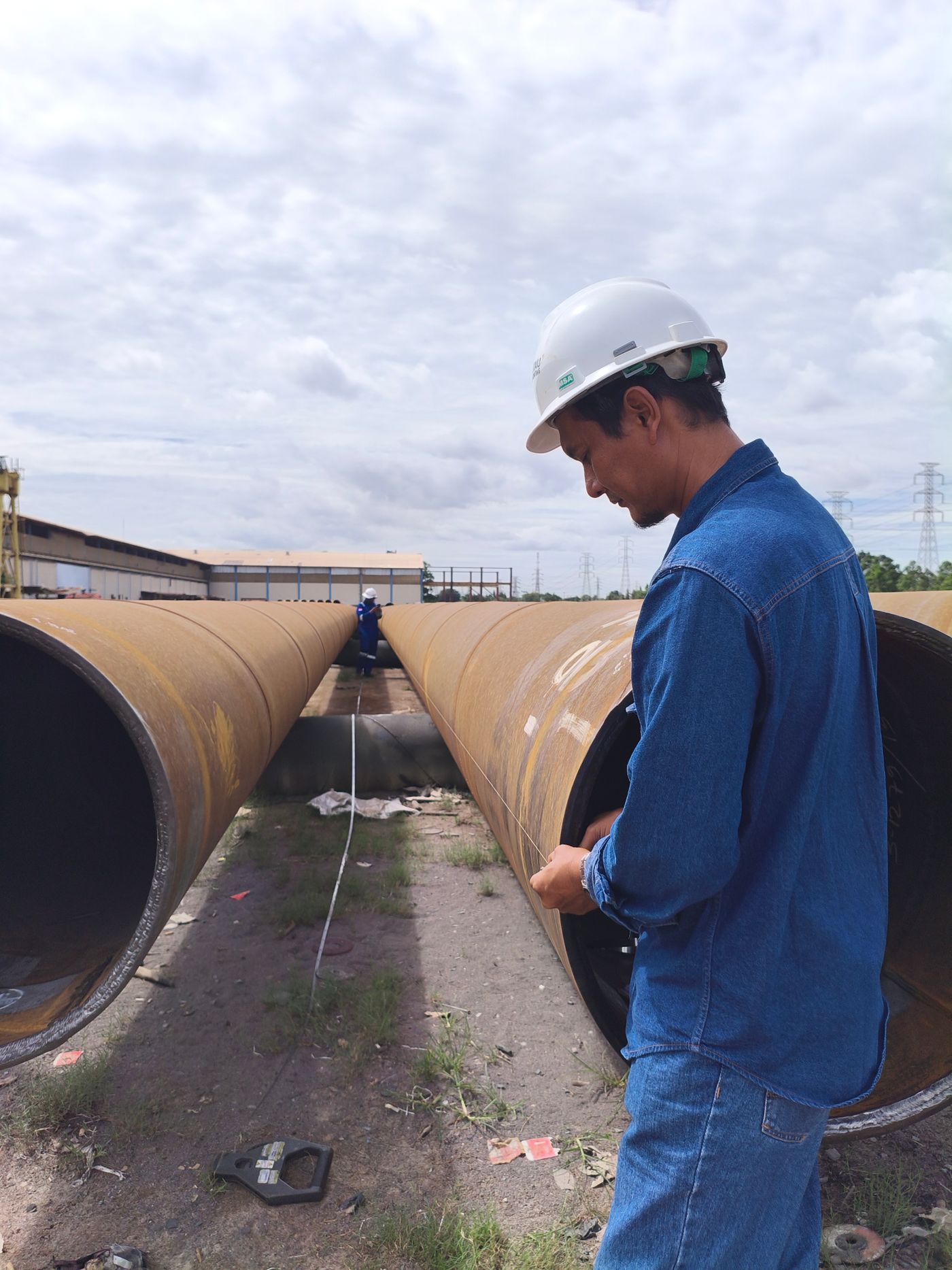 A worker in a hard hat inspecting large pipes at a pipeline yard, with a second worker further down the line