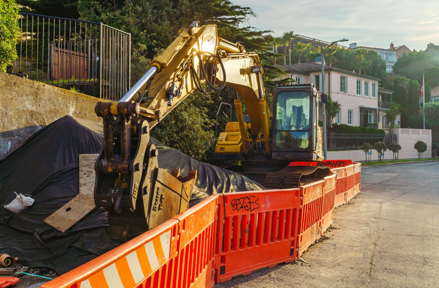 A yellow excavator on a residential street construction site behind orange safety barriers, with houses behind