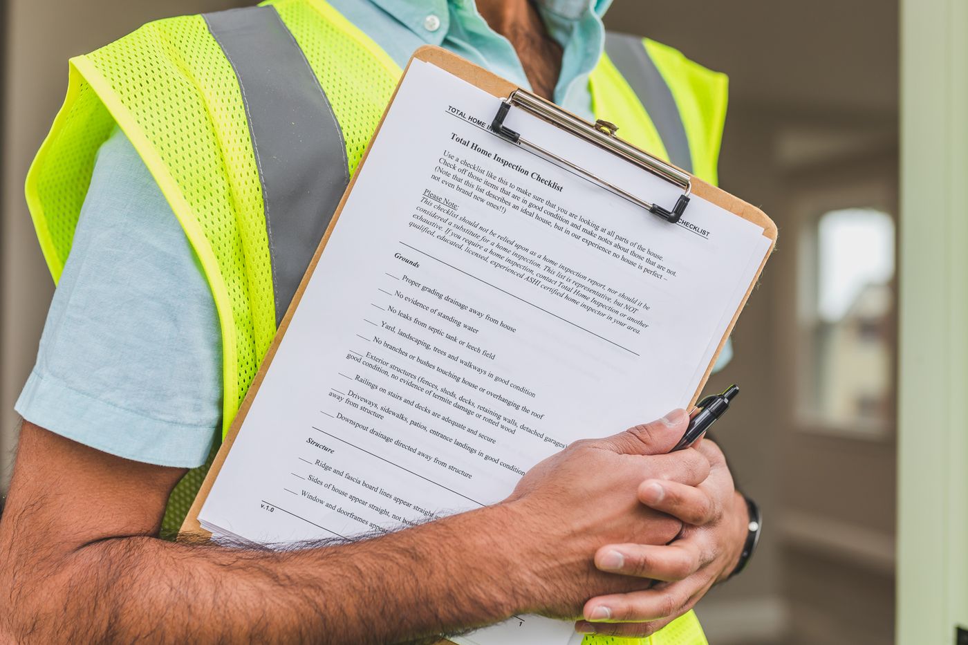 An inspector in a yellow safety vest holding a clipboard with a home inspection checklist and a pen