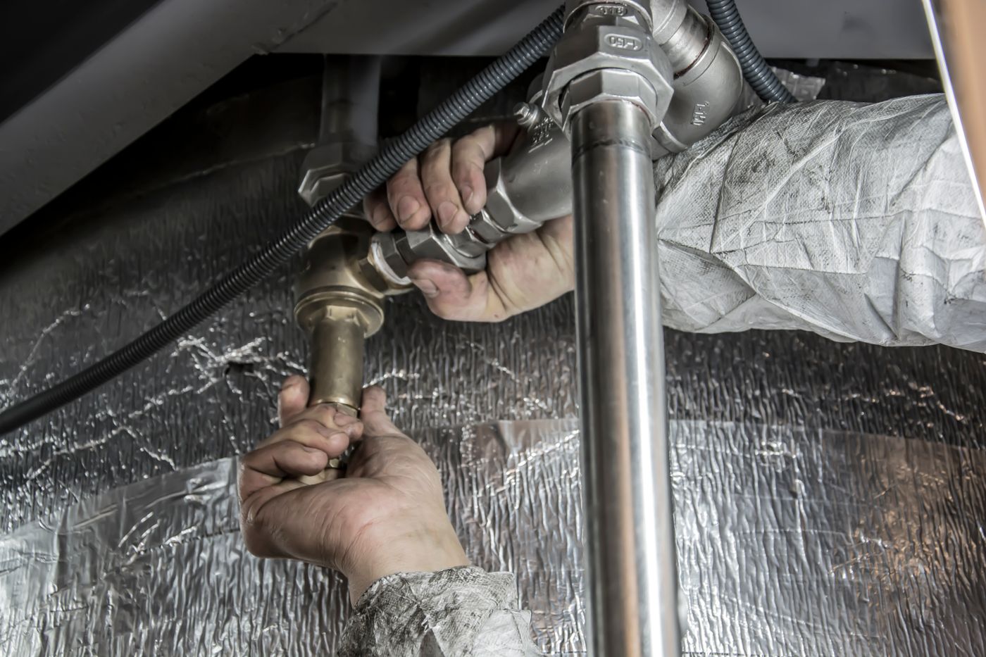 Close-up of a plumber's hands connecting brass and stainless steel pipe fittings under insulation