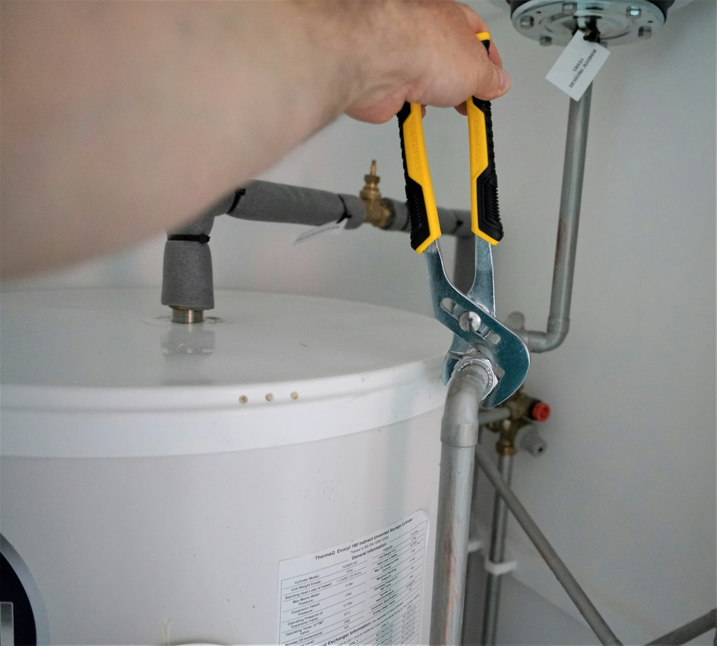 A technician using pliers to work on the pipe connections atop a white hot water storage cylinder